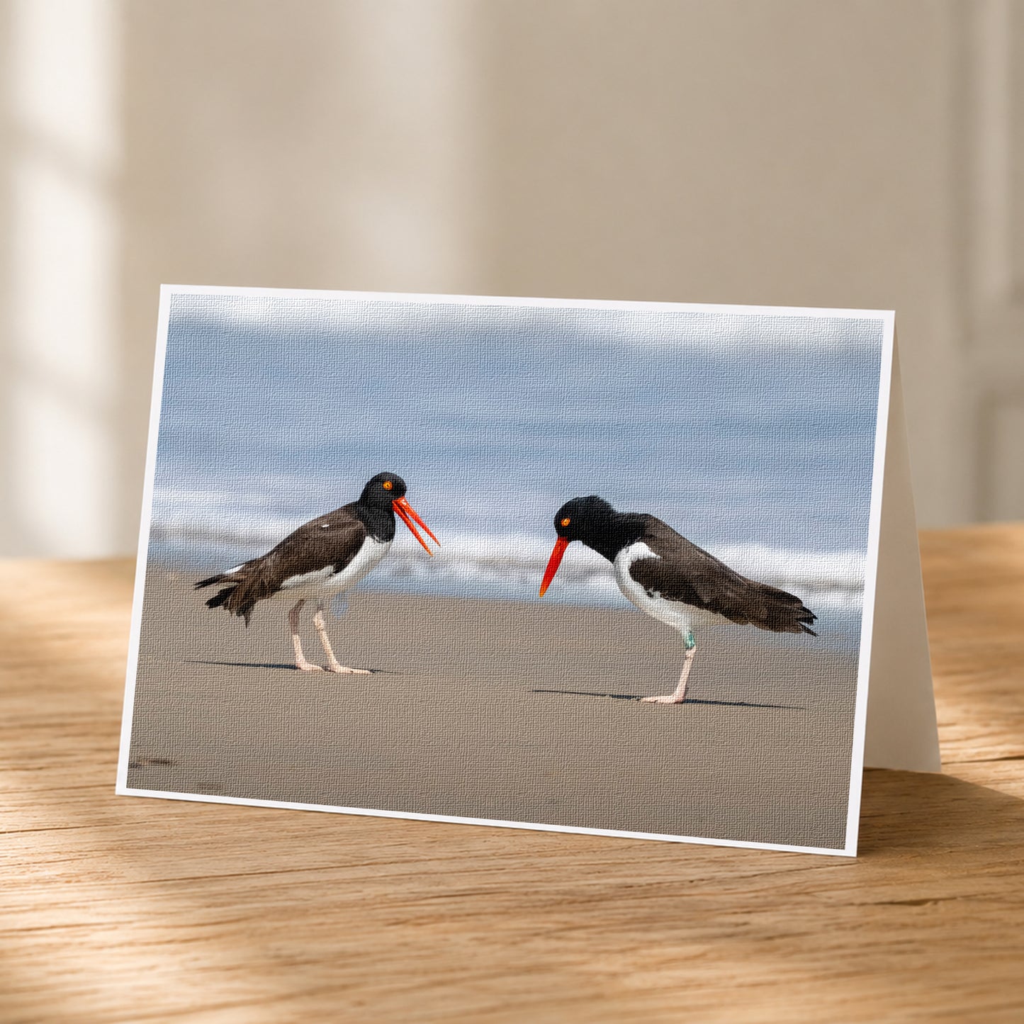 A greeting card featuring two black and white birds with orange beaks standing on a sandy beach, facing each other.