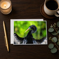 a close-up of a black bird with a yellow beak, possibly a heron or egret, on a wooden surface. There is also a candle, a pen, and a cup of coffee nearby.