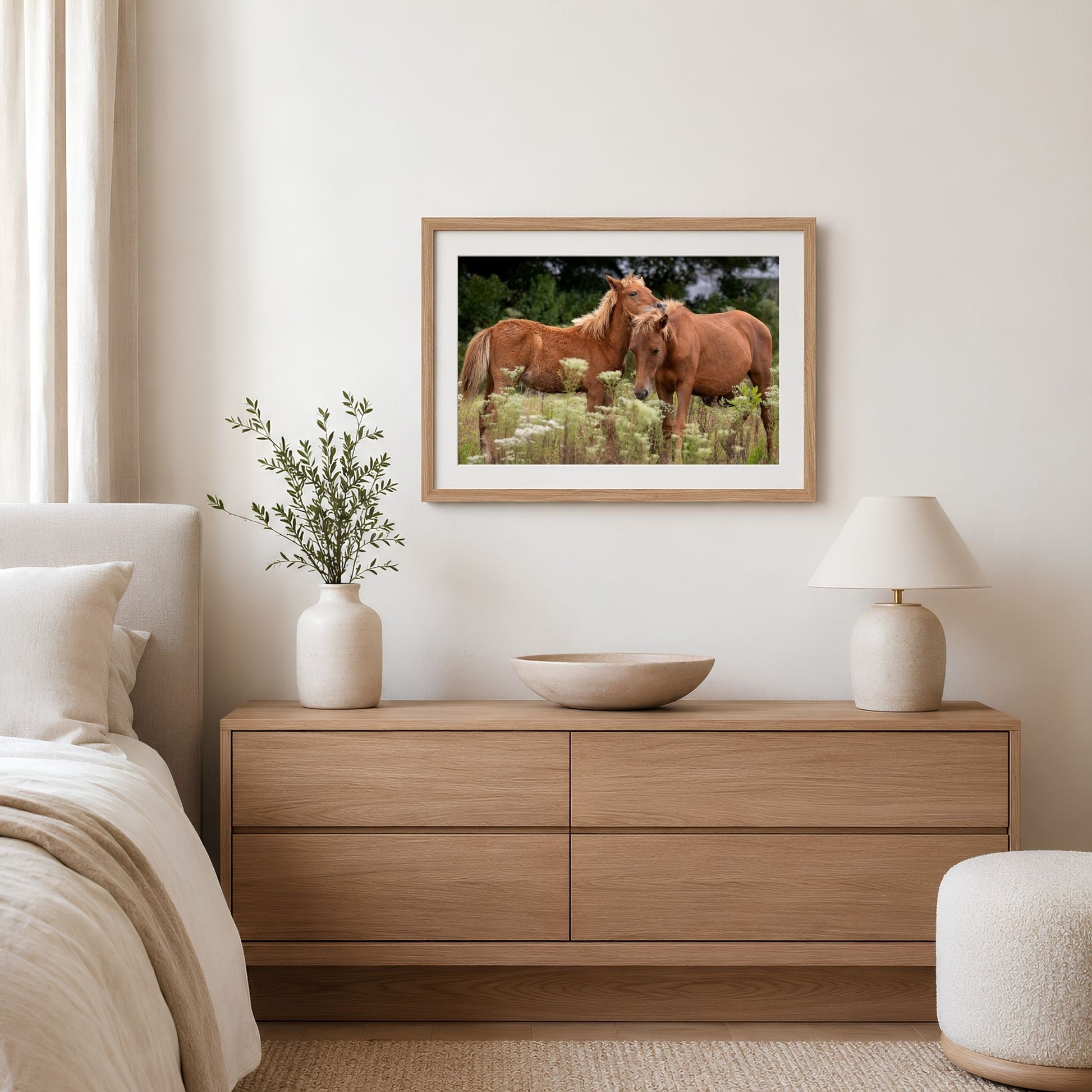 a bedroom with a wooden dresser, a framed picture of two horses, a lamp, and a plant.