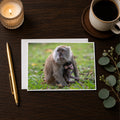 a card with a photograph of a monkey sitting on the grass, accompanied by a candle, a pen, and a cup of coffee on a wooden surface.