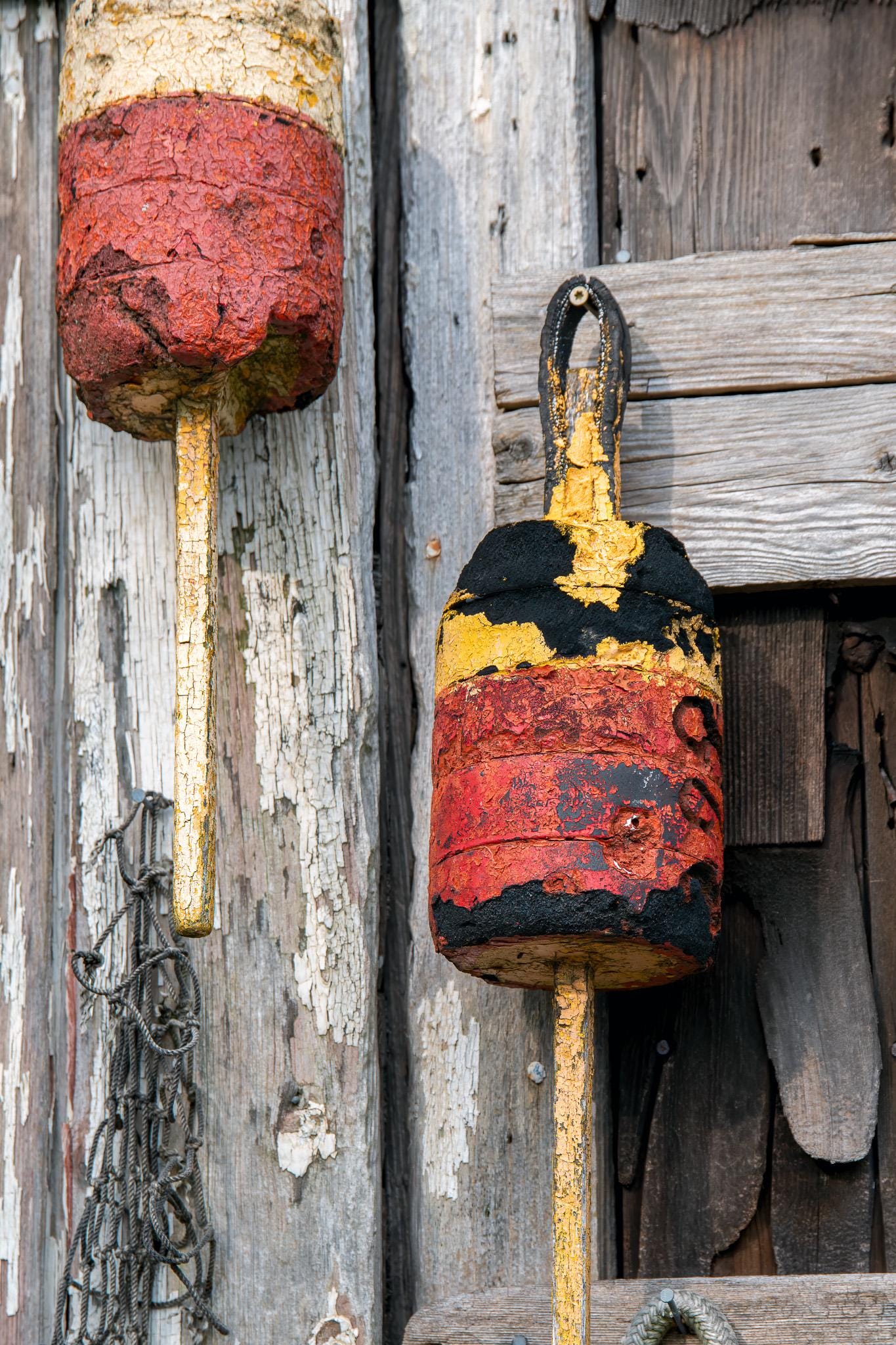 Weathered lobster buoys hanging on an aged wooden wall in Rockport, Massachusetts, with peeling paint, rope, cracked red and yellow fishing floats, and rustic New England coastal texture photographed by Linda Nee.