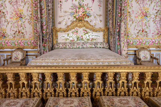 Marie Antoinette’s floral royal bedchamber at the Palace of Versailles, featuring an ornate canopy bed, gilded gold railing, pink floral fabric walls, matching chairs, and French palace decor.