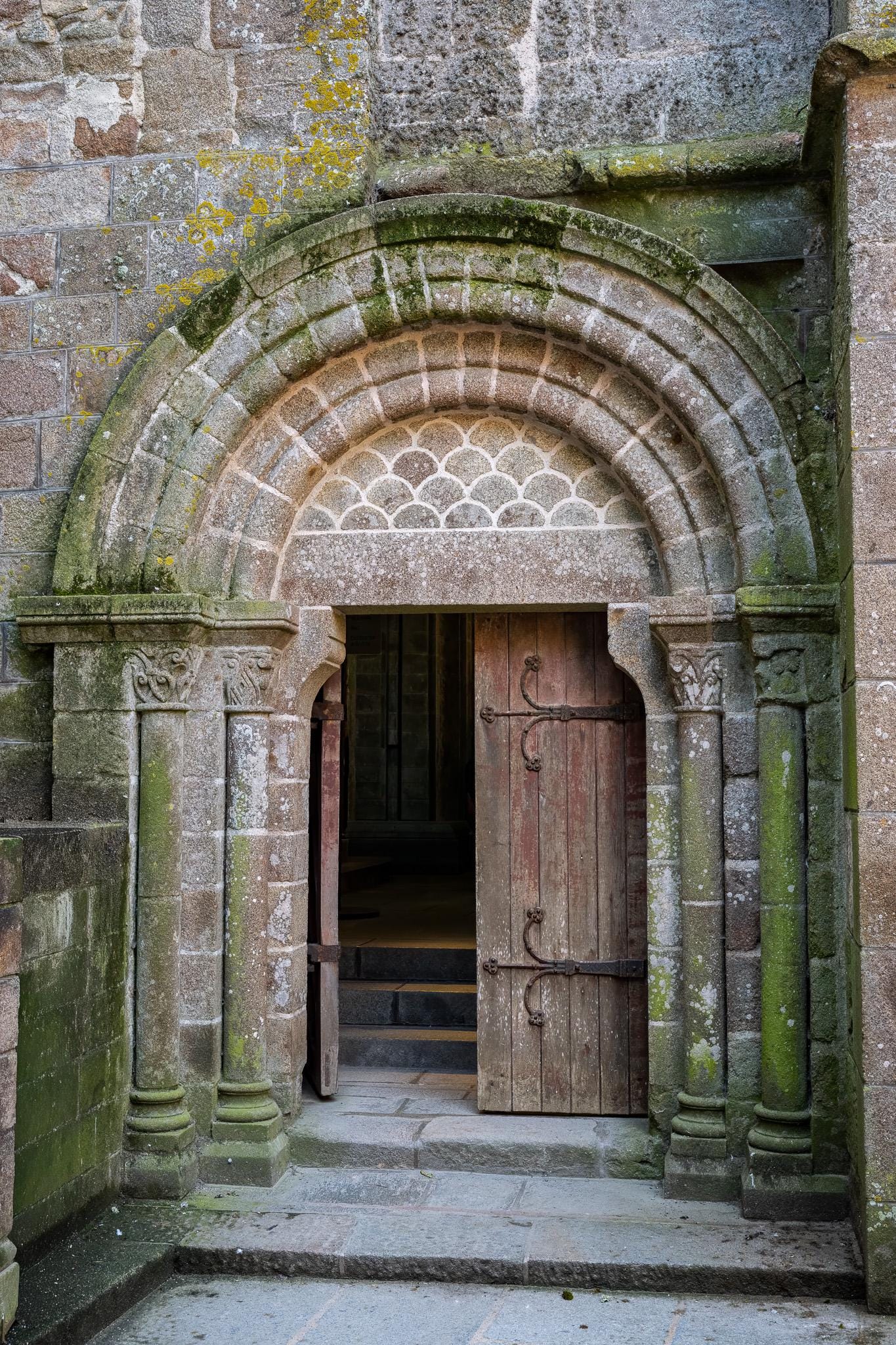 Open Romanesque stone doorway at Mont Saint-Michel Abbey in Normandy, France, with a weathered wooden door, moss-covered medieval stonework, carved columns, and steps leading into the abbey interior.