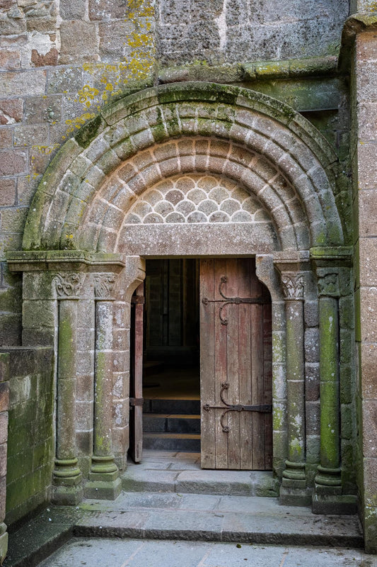 Open Romanesque stone doorway at Mont Saint-Michel Abbey in Normandy, France, with a weathered wooden door, moss-covered medieval stonework, carved columns, and steps leading into the abbey interior.