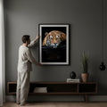 A man is hanging a framed photograph of a tiger on a wall in a living room.