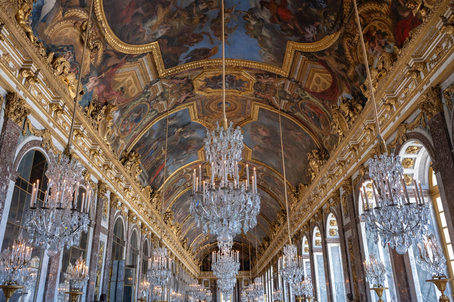Palace of Versailles Hall of Mirrors interior with crystal chandeliers, gilded trim, painted ceiling murals, arched windows, and ornate French Baroque architecture.