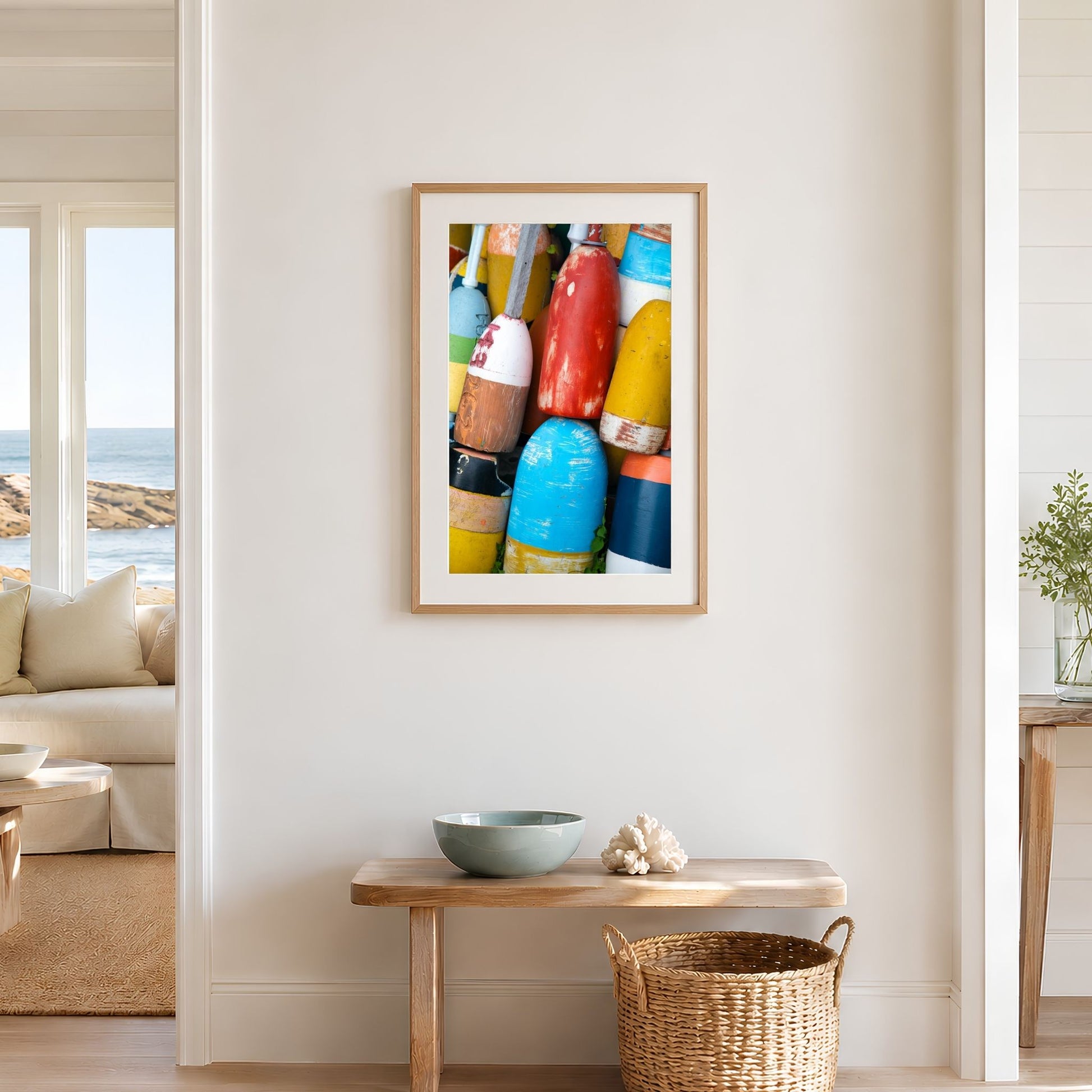 Framed vertical colorful lobster buoy photography print in a natural oak frame on a warm white coastal wall above a wooden bench and woven basket