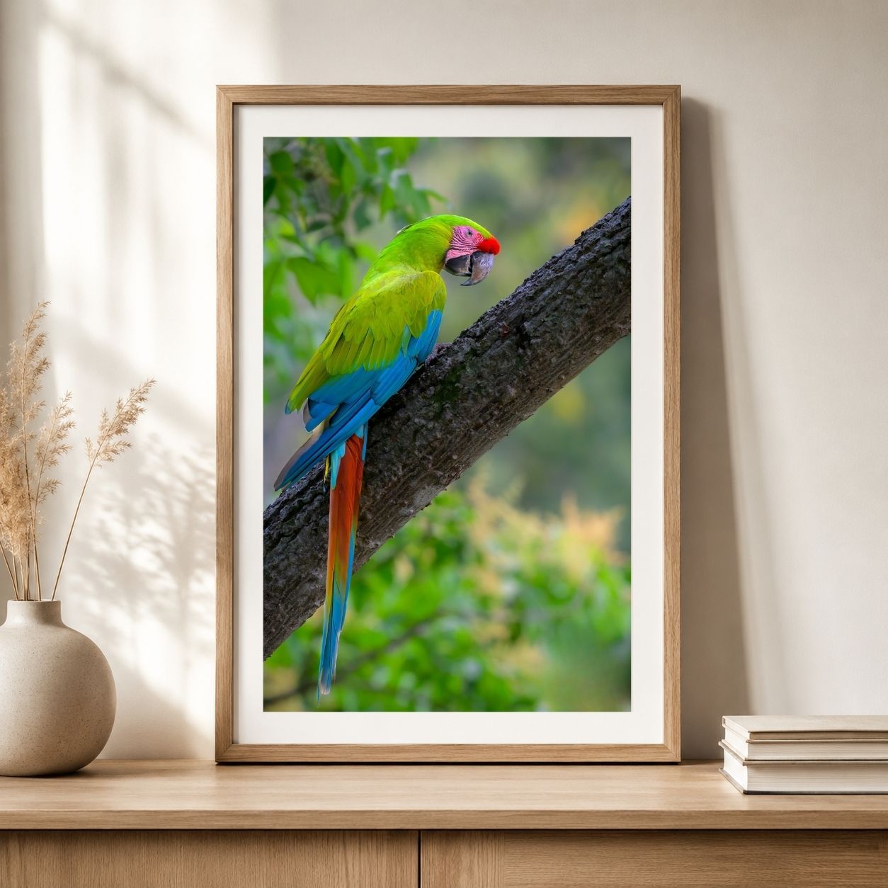 Framed fine art print of a great green macaw perched on a tree branch, displayed in a light wood frame on a neutral console table.