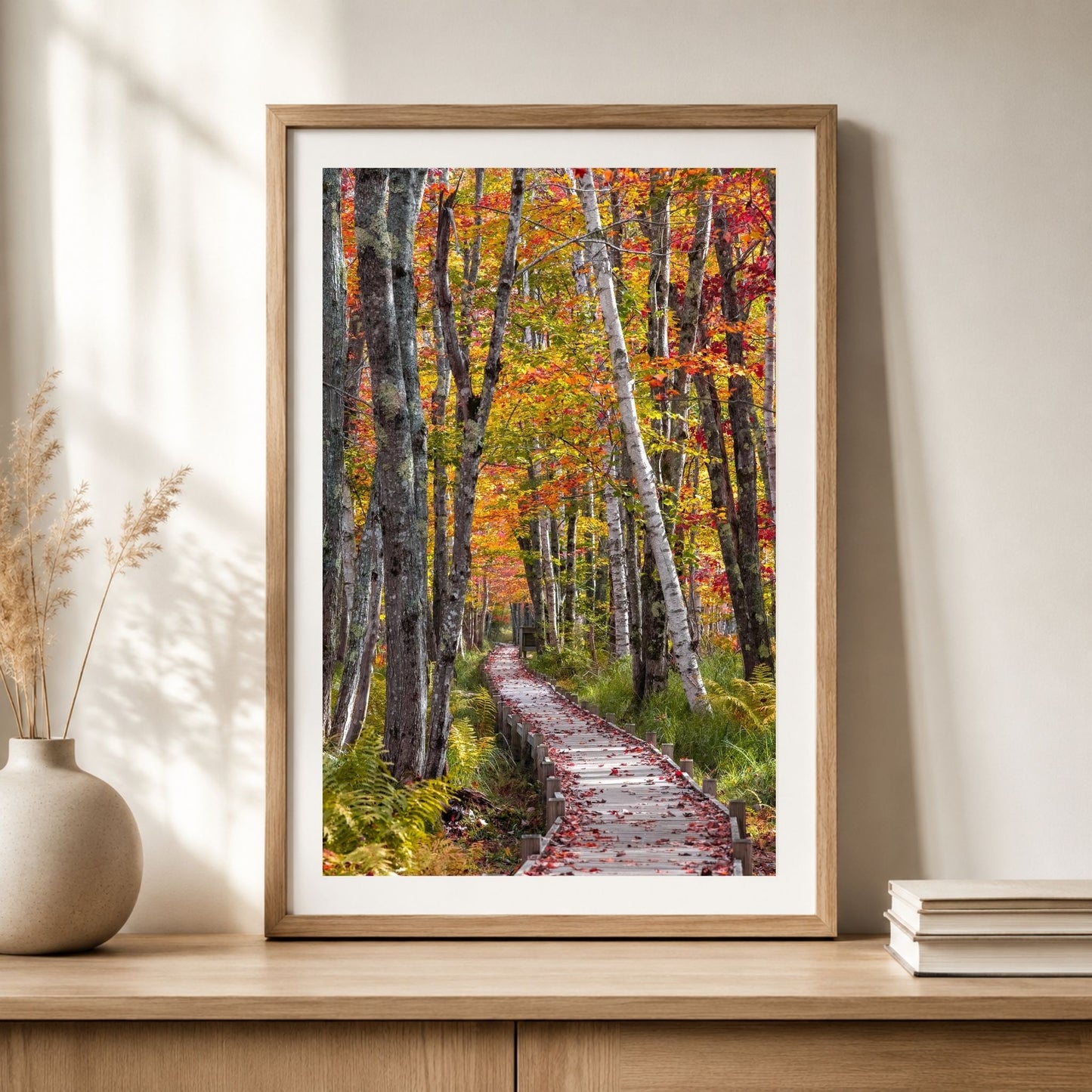 Framed vertical Acadia autumn forest boardwalk photography print in a natural oak frame with white mat, displayed on a light wood console with neutral home decor