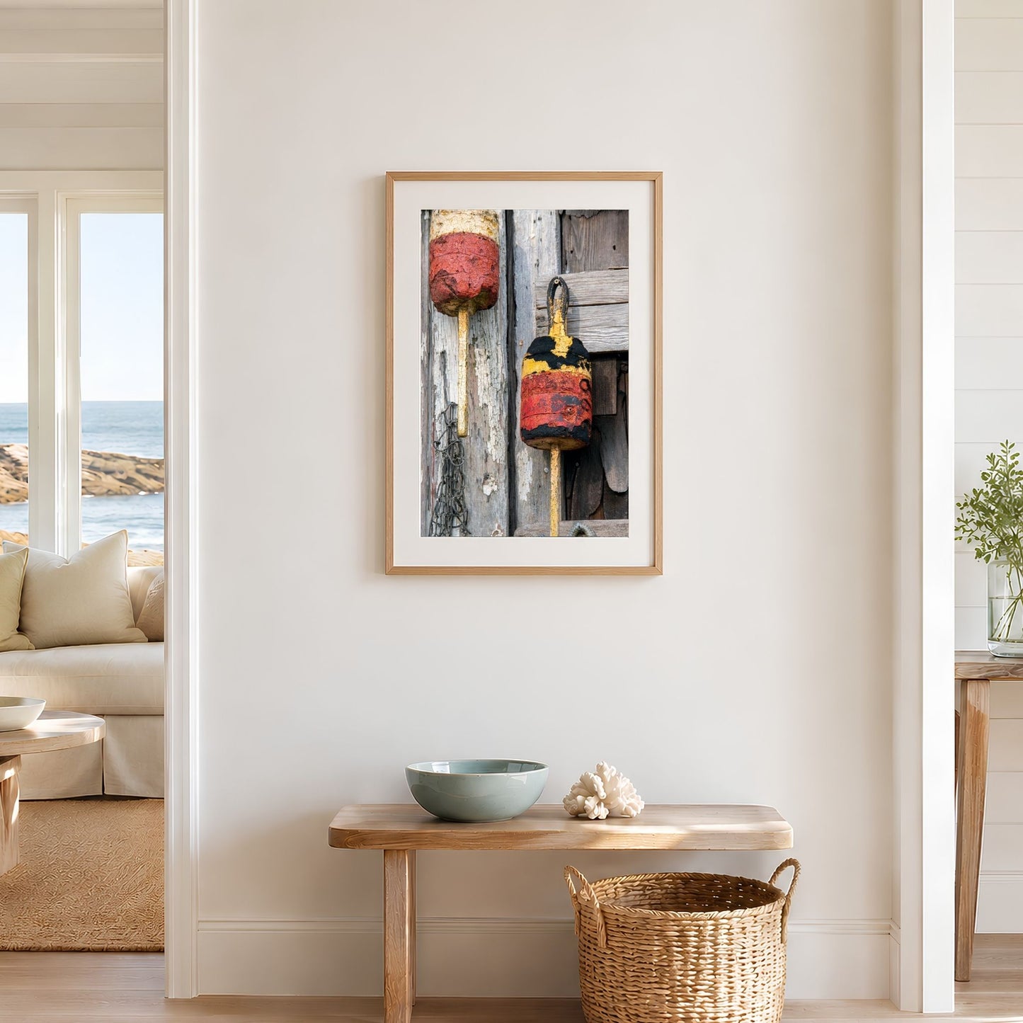 Framed vertical lobster buoy photography print in a natural oak frame on a warm white beach house wall above a wooden bench and woven basket