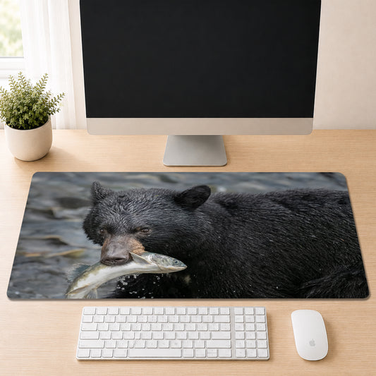 Black bear standing in a river with a freshly caught salmon in its mouth, shown as a desk mat design on a wood desk