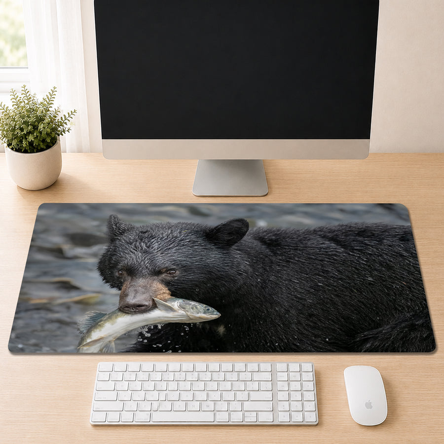Black bear standing in a river with a freshly caught salmon in its mouth, shown as a desk mat design on a wood desk