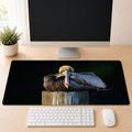 a computer monitor, keyboard, and mouse on a desk, with a large mouse pad displaying a close-up of a pelican resting on a wooden post.