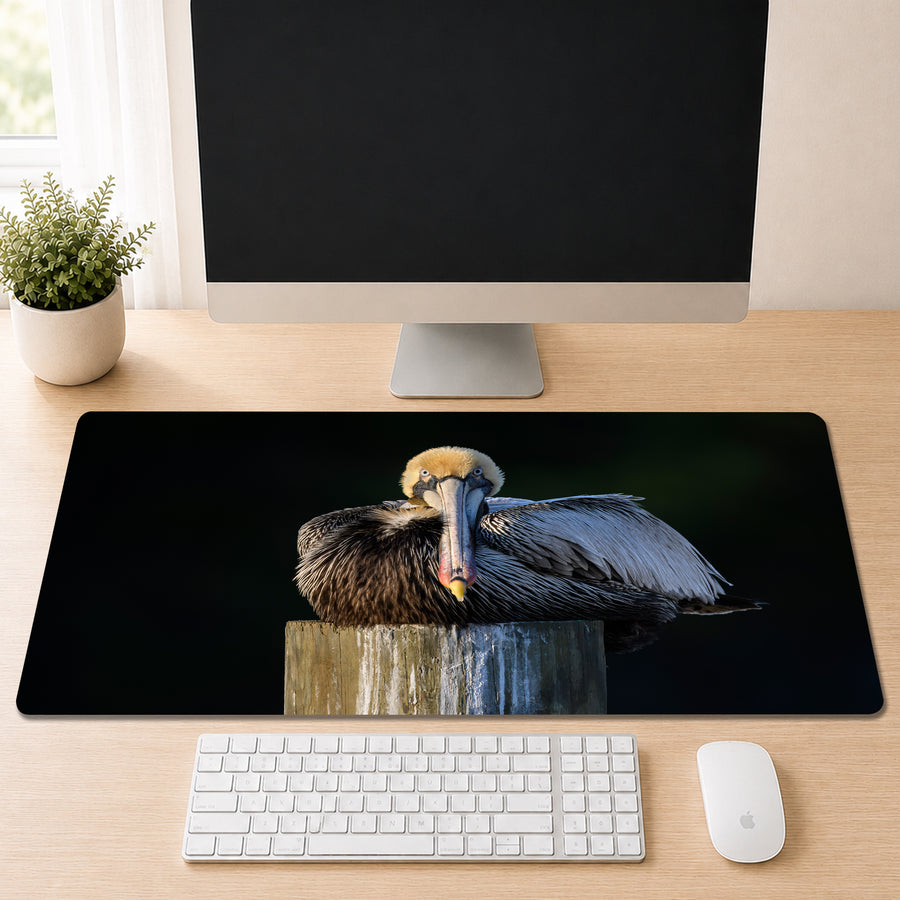 a computer monitor, keyboard, and mouse on a desk, with a large mouse pad displaying a close-up of a pelican resting on a wooden post.