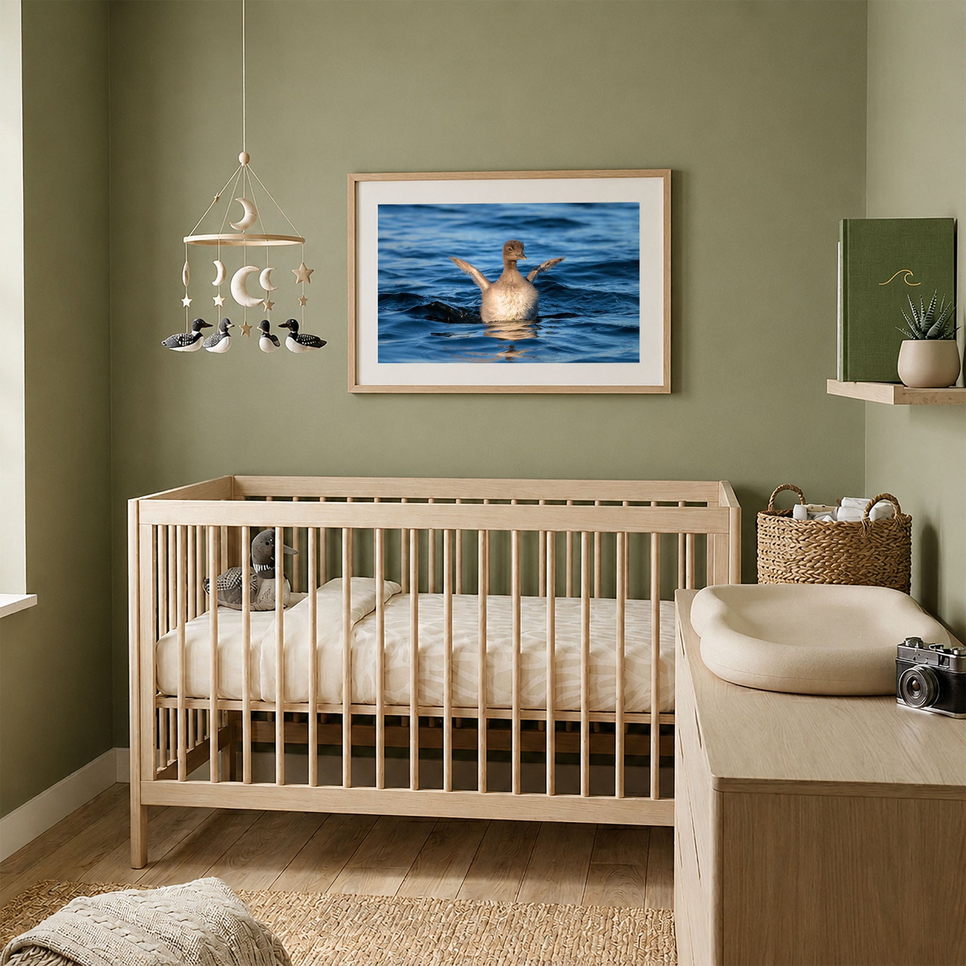 Framed baby loon chick photograph displayed above a natural wood crib in a cozy sage green nursery.