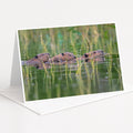 A greeting card featuring three beavers swimming in a body of water surrounded by tall grass.