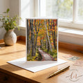 A greeting card featuring a scenic autumn forest path is displayed on a wooden desk, with a pen and postage stamps nearby.