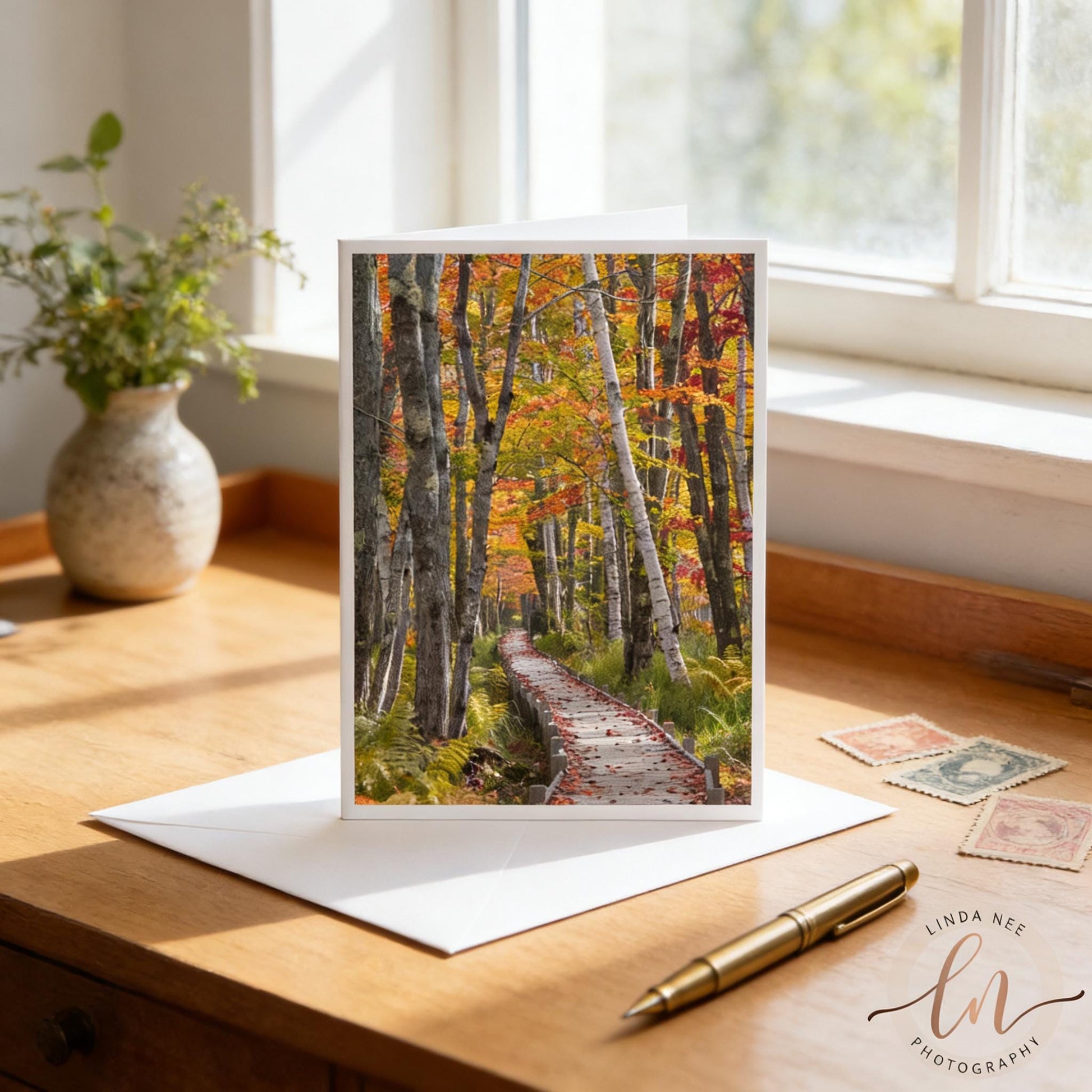 A greeting card featuring a scenic autumn forest path is displayed on a wooden desk, with a pen and postage stamps nearby.