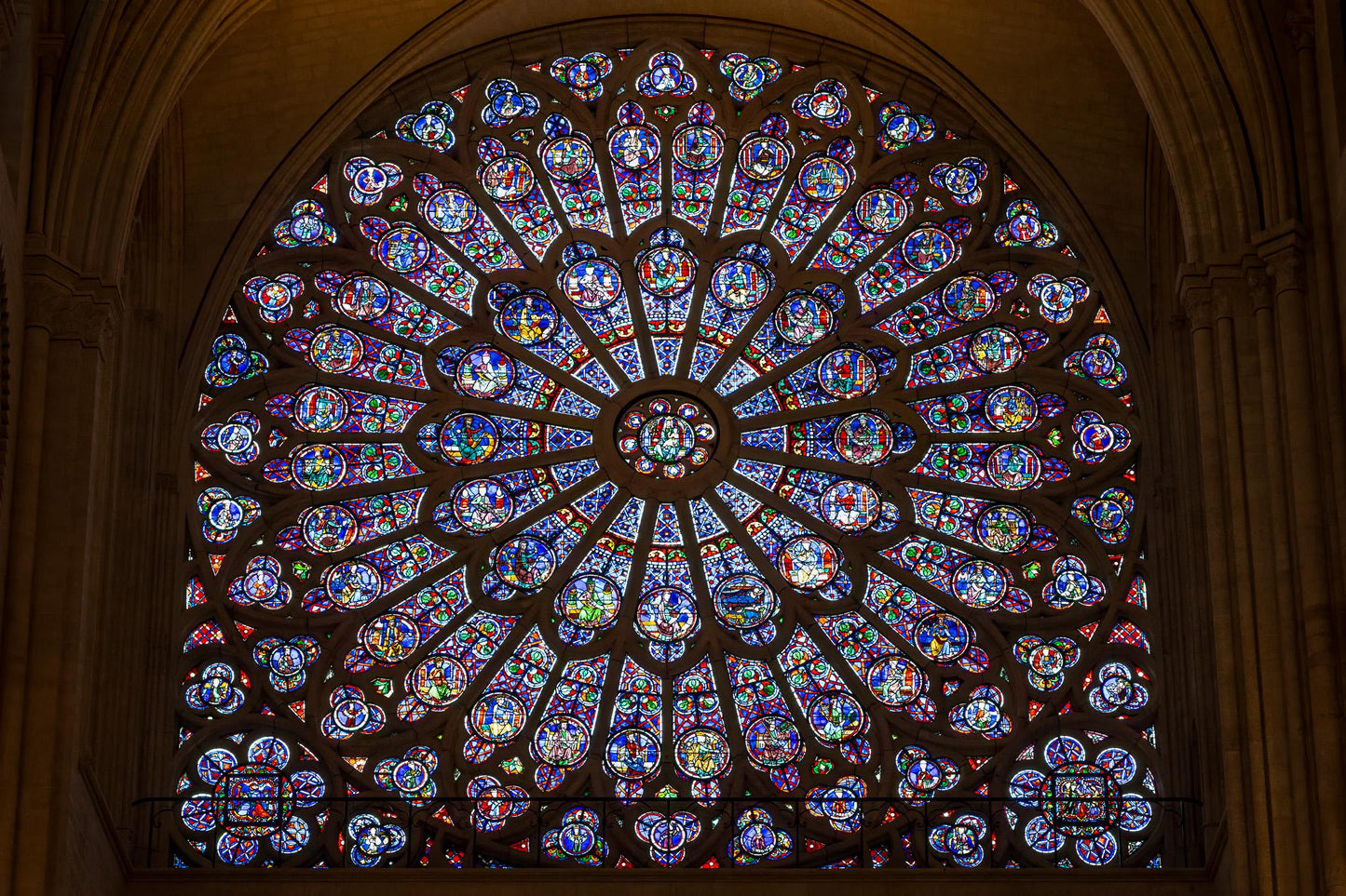 Colorful rose window inside Notre-Dame de Paris, framed by dark Gothic stone arches and glowing stained glass panels in red, blue, green, and gold.