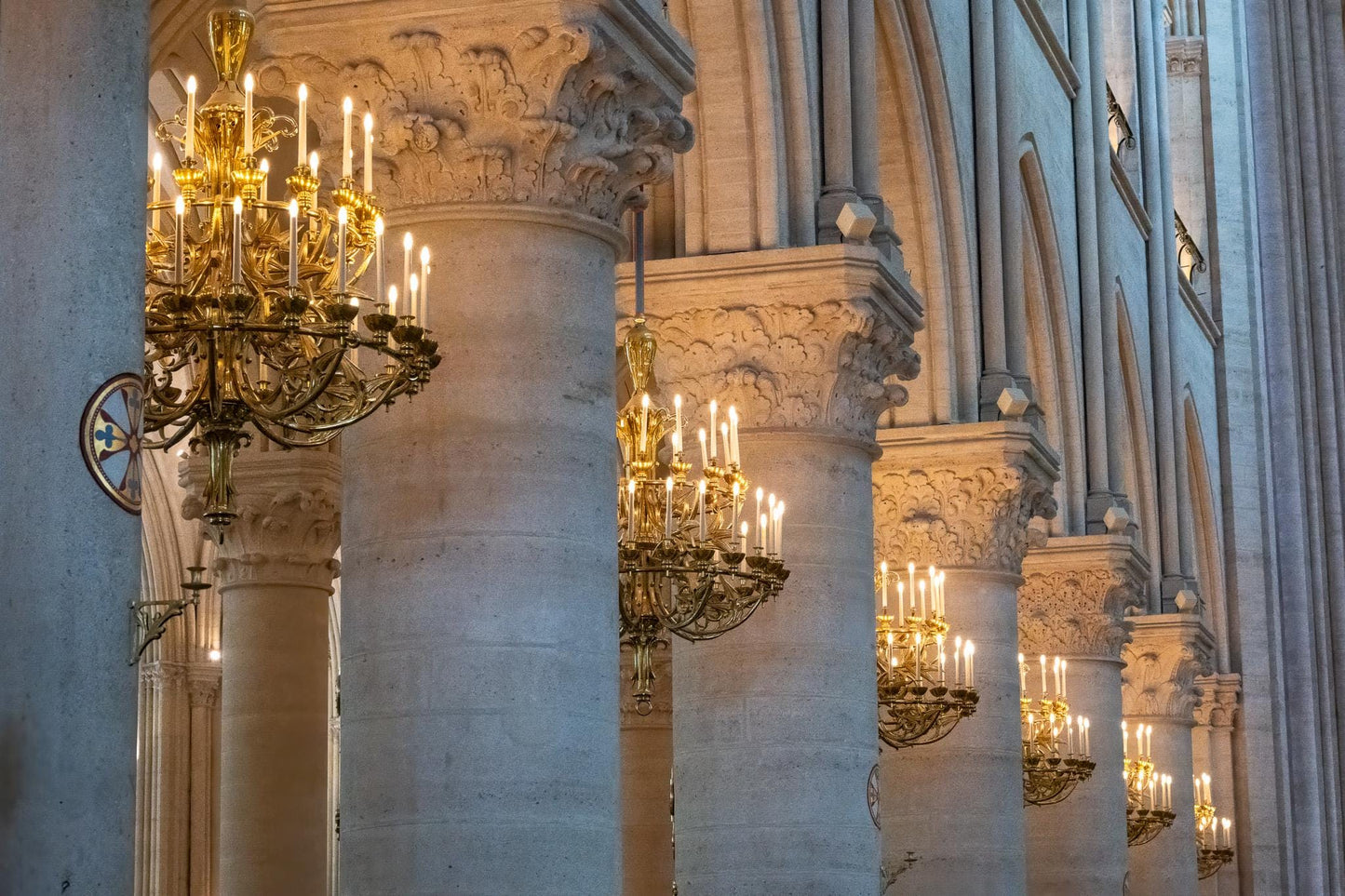 Restored interior of Notre-Dame Cathedral in Paris with pale limestone columns, ornate carved capitals, and glowing gold chandeliers receding through the historic Gothic nave.